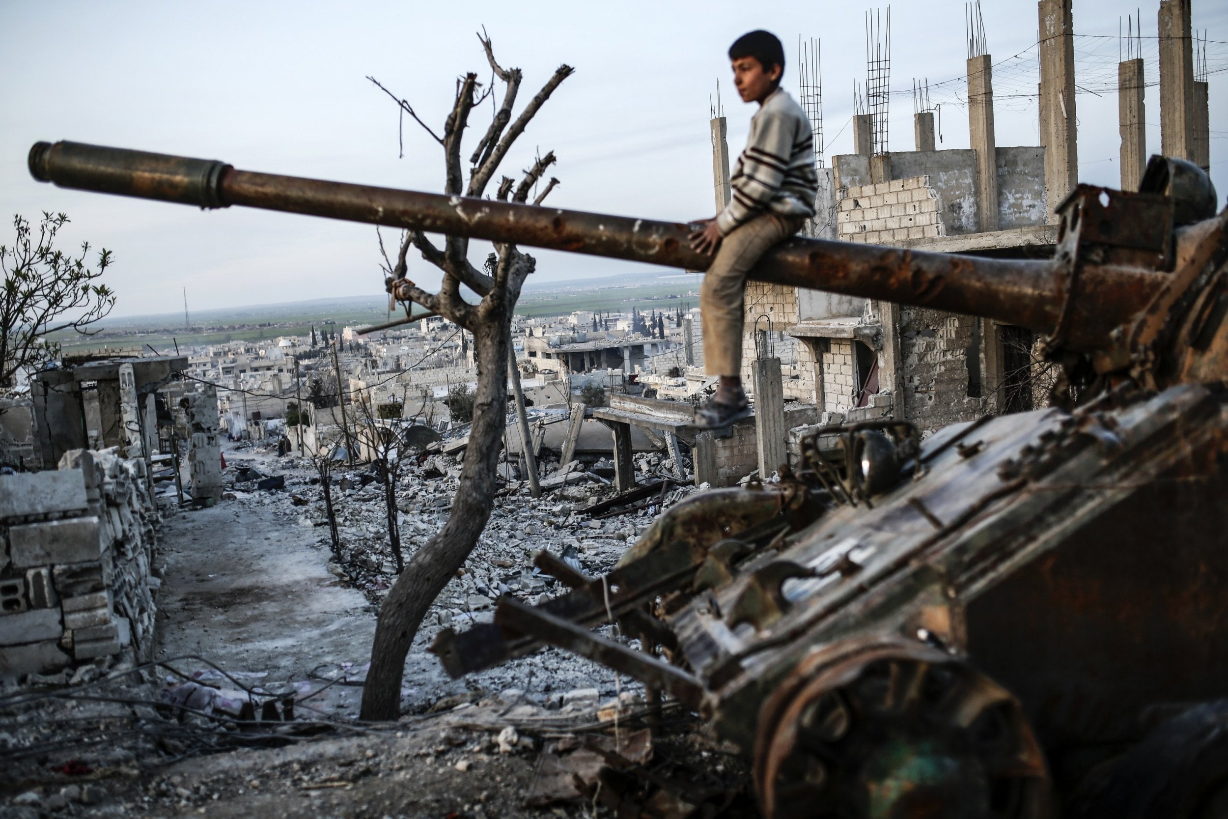 A boy sits on a rusted tank barrel amidst a war-torn landscape of ruined buildings and debris, with barren trees in the foreground.