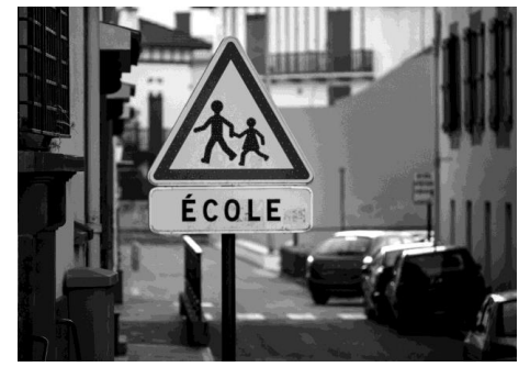 Triangular school crossing sign with two figures holding hands, labelled "École," on an urban street with parked cars and buildings in the background.