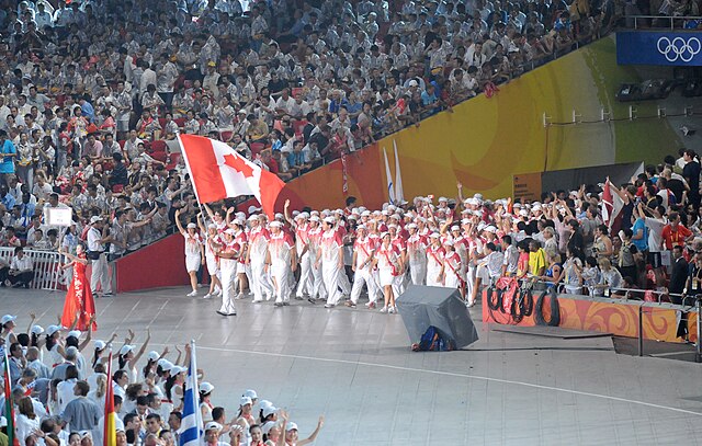 Athletes in white uniforms with red accents parade with a large Canadian flag during an Olympic opening ceremony, surrounded by a cheering crowd.