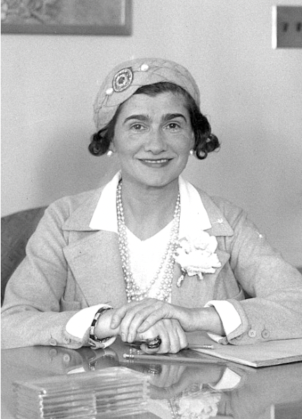 Coco Chanel seated at a desk, wearing a stylish hat and suit with a flower brooch and pearl necklace, smiling directly at the camera.