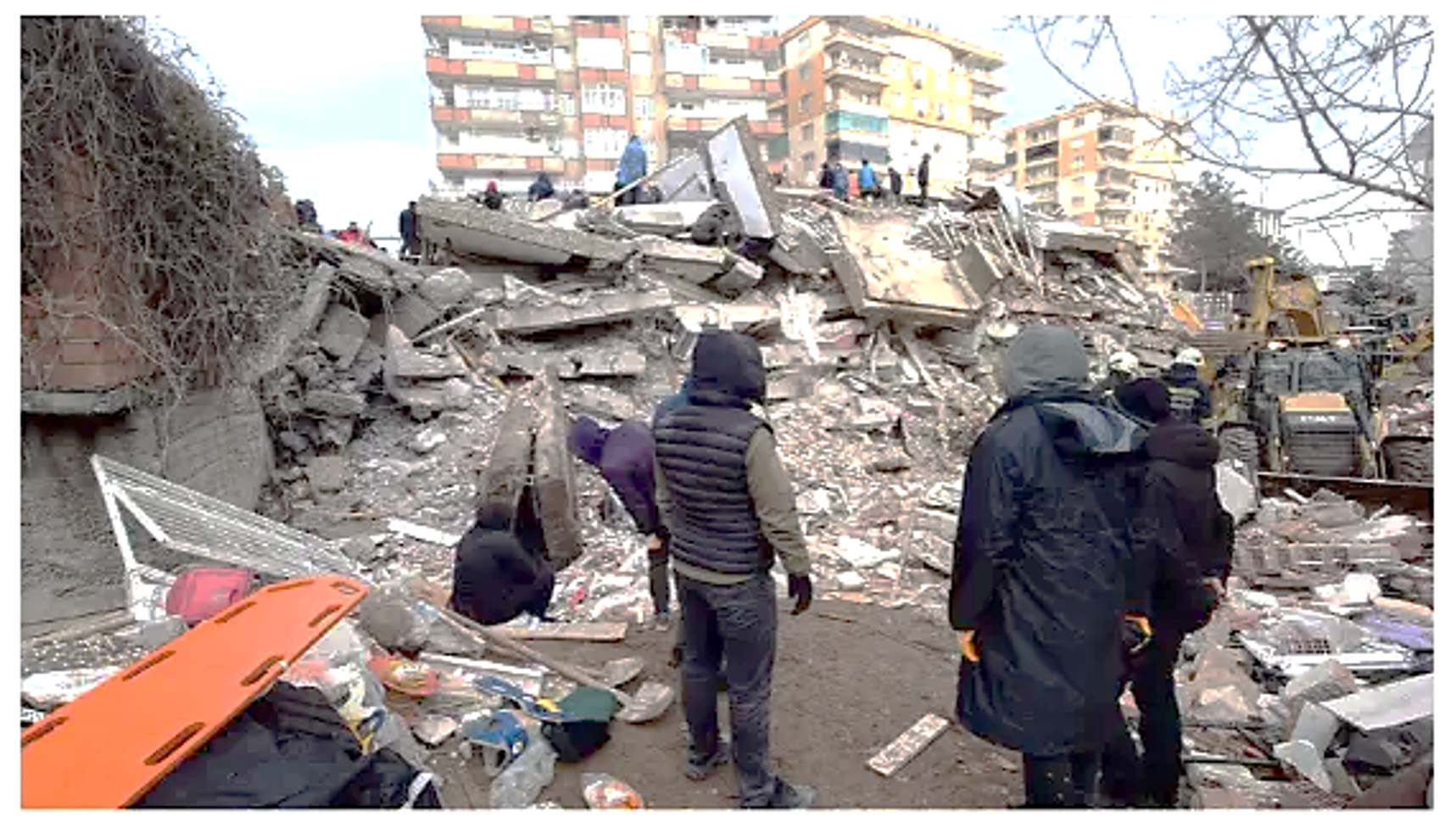 People stand amid collapsed building rubble, likely from an earthquake, with debris scattered and rescue workers visible in the background.