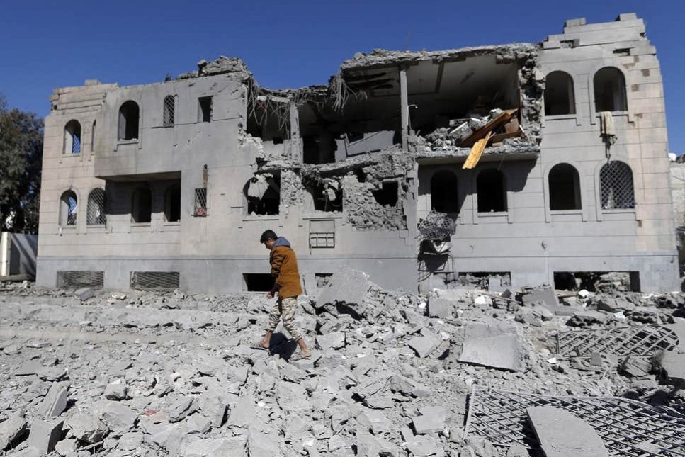 Man walking on debris in front of a heavily damaged building with partially collapsed walls and broken windows, under a clear blue sky.