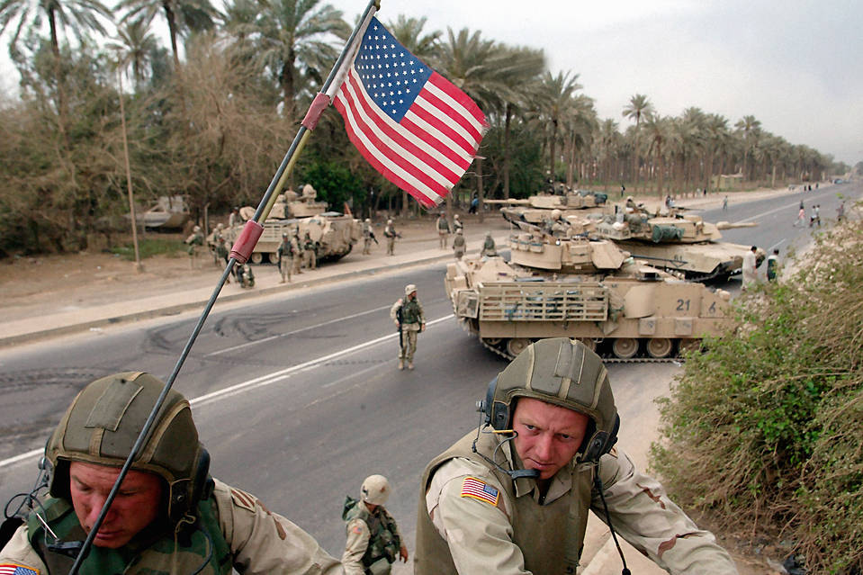 Two soldiers in helmets atop a vehicle with an American flag, palm trees, tanks, and troops on a road in the background.
