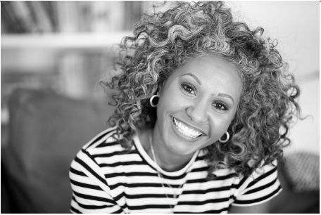 Smiling woman with curly hair wearing a striped shirt and earrings, seated indoors with books blurred in the background, captured in black and white.