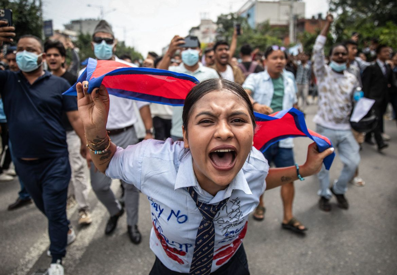 A passionate protester holds a flag, shouting amidst a crowd on the street. Others behind display similar fervour, some wearing face masks.