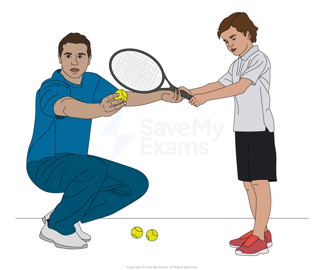 A tennis coach in a tracksuit instructs a young boy holding a racket on an indoor court. Numerous tennis balls are scattered on the floor.