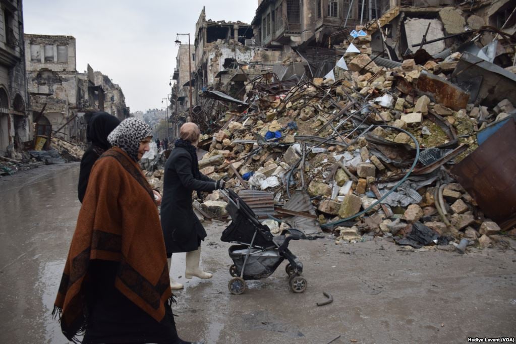 Three people walk past a large pile of rubble and debris, including bricks and metal, on a wet, overcast street with damaged buildings.