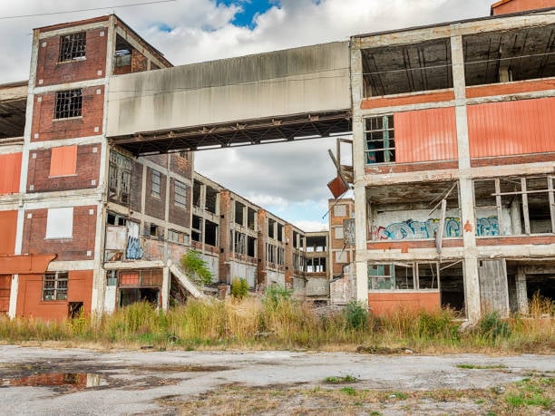 Abandoned industrial building with weathered brick and metal, shattered windows, graffiti, and overgrown vegetation under a cloudy sky.