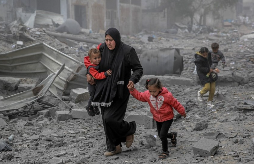 A woman carrying a child and holding a girl's hand walks through rubble and debris, with more children in the background, in an area affected by conflict.