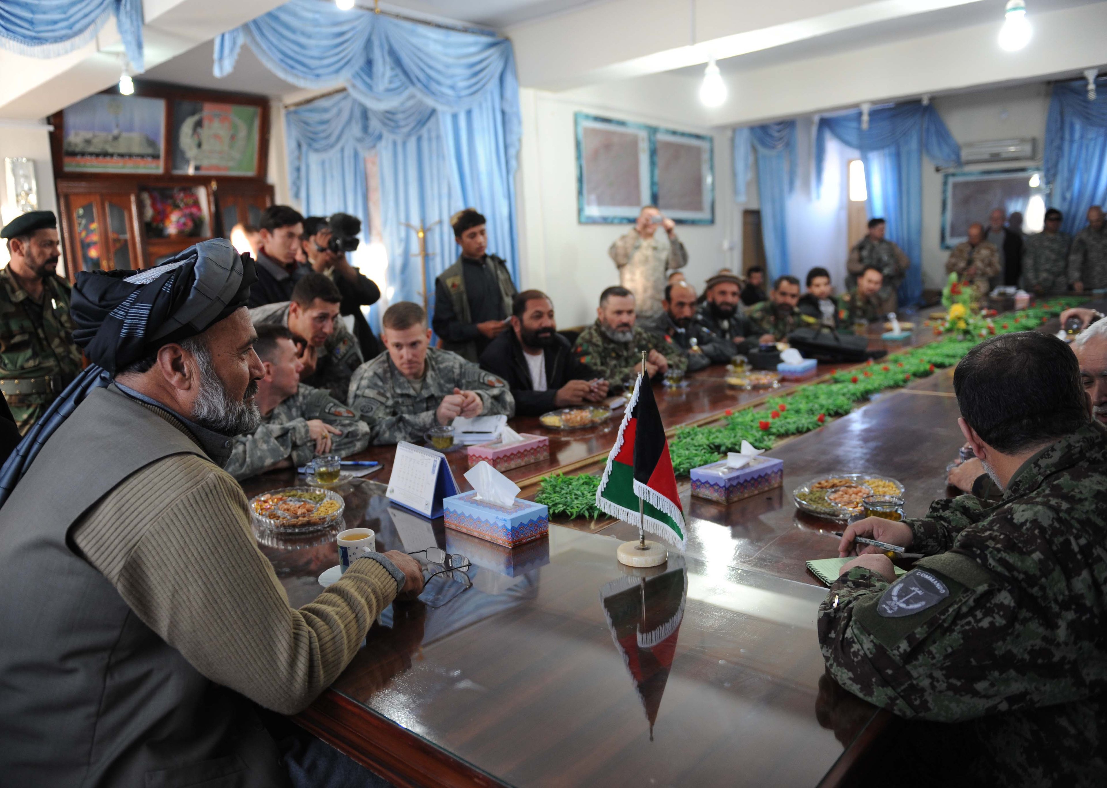 Military and civilian leaders seated around a long table during a meeting, with Afghan flag and refreshments in a room with blue curtains.