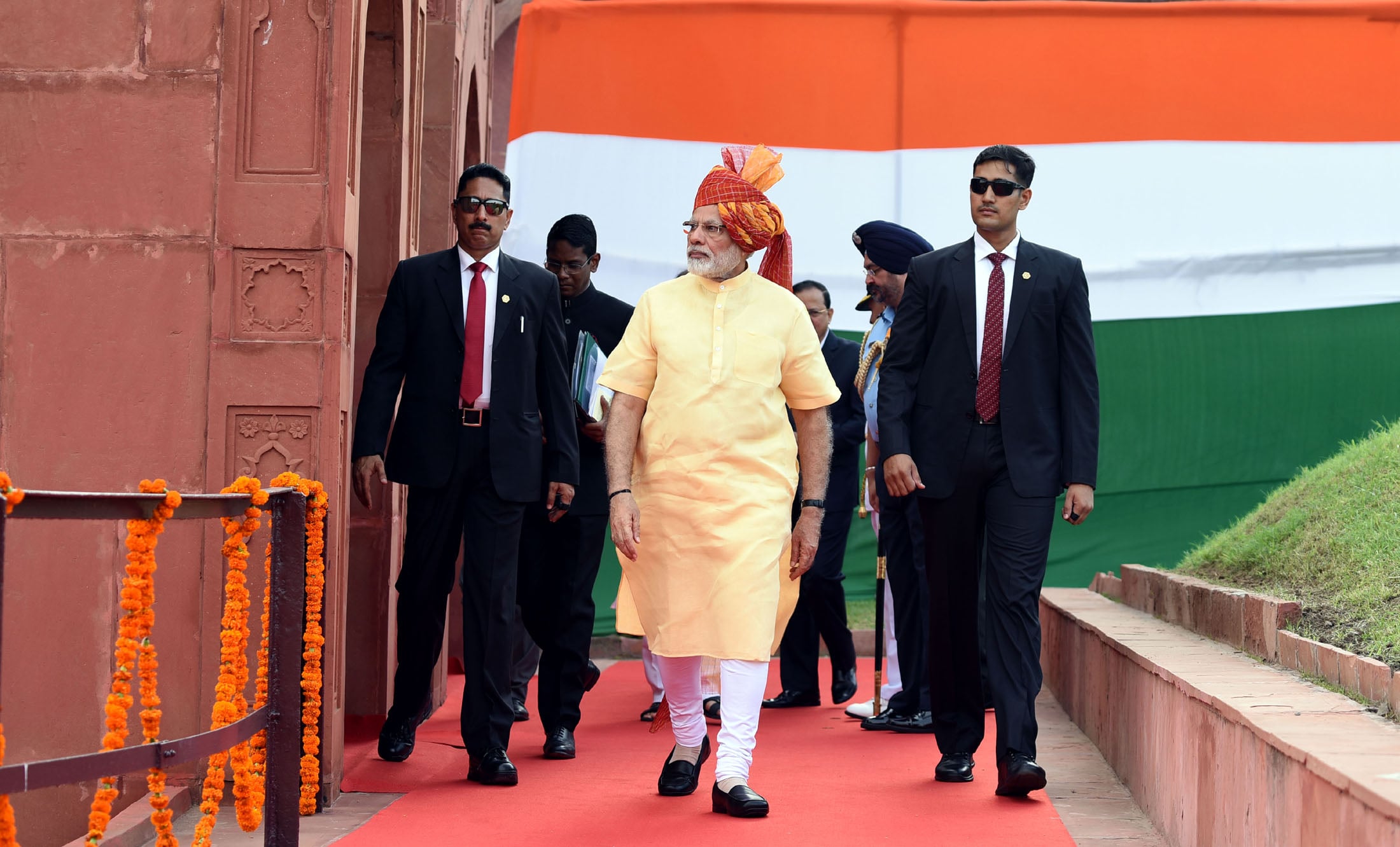 Man in traditional attire walks on a red carpet flanked by security personnel. Large Indian flag in background, building adorned with orange flowers.
