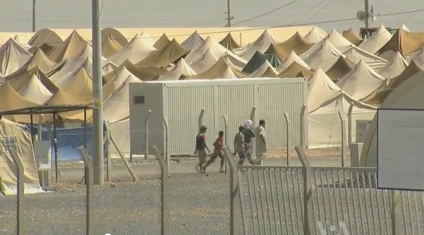 Rows of beige tents in a refugee camp with a chain-link fence in the foreground and several people walking along a gravel path.