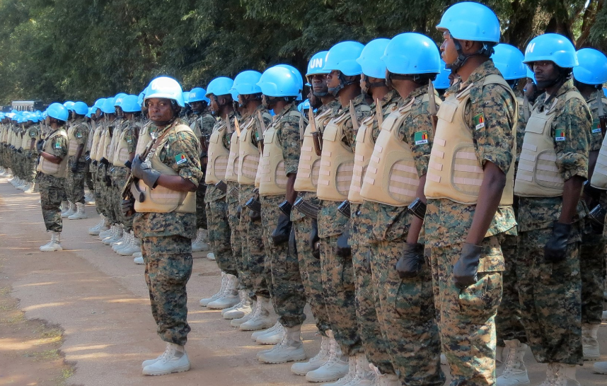 Soldiers in camouflage uniforms and blue helmets stand in formation on a paved road, with trees in the background, indicating a peacekeeping mission.
