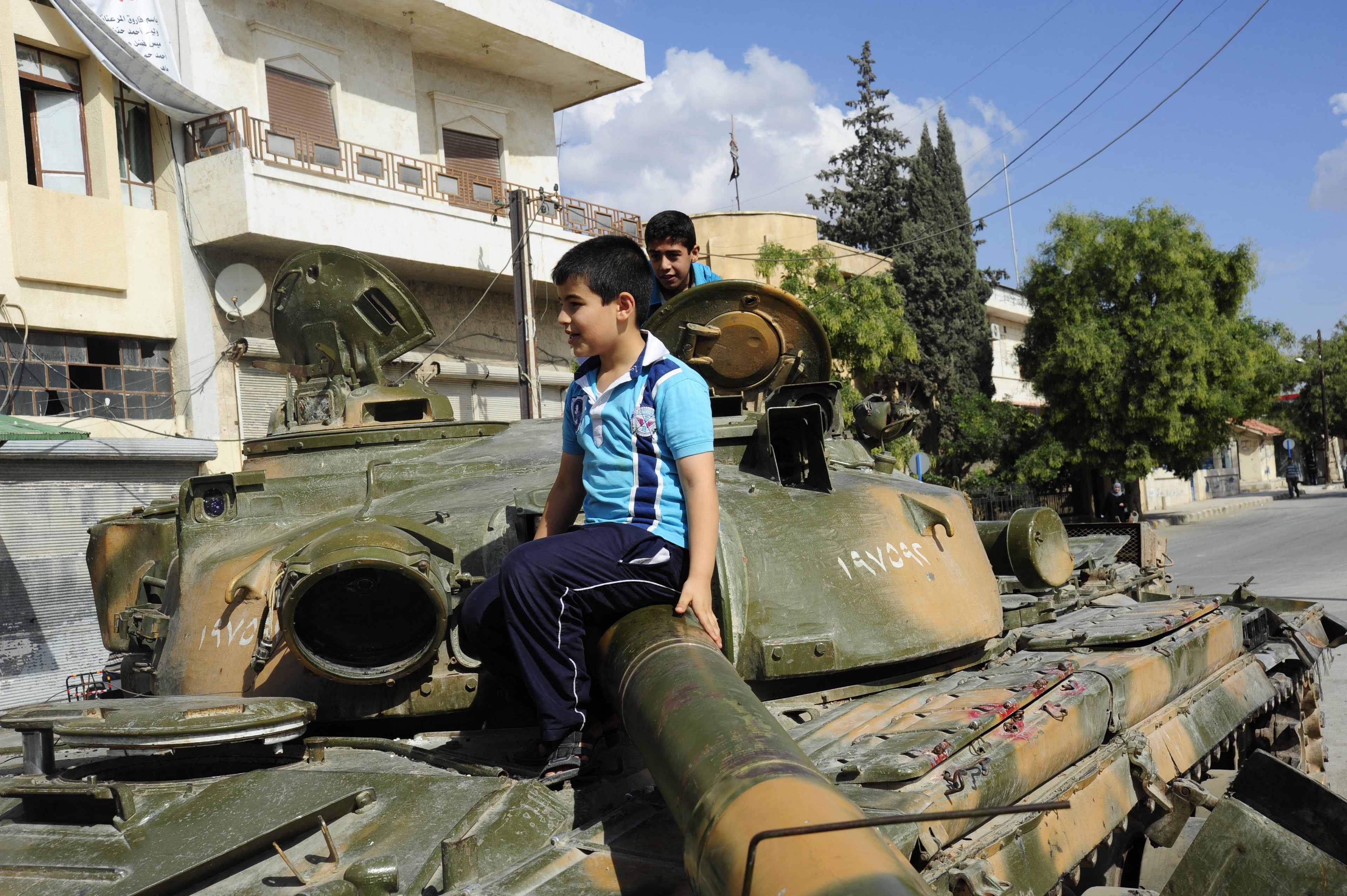 Two boys sit and stand on a rusted military tank parked on a street in an urban area, with buildings and trees in the background.