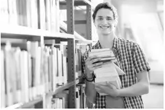 Young man smiling, holding a stack of books in a library aisle. Shelves filled with books are visible, and soft light filters through a window.