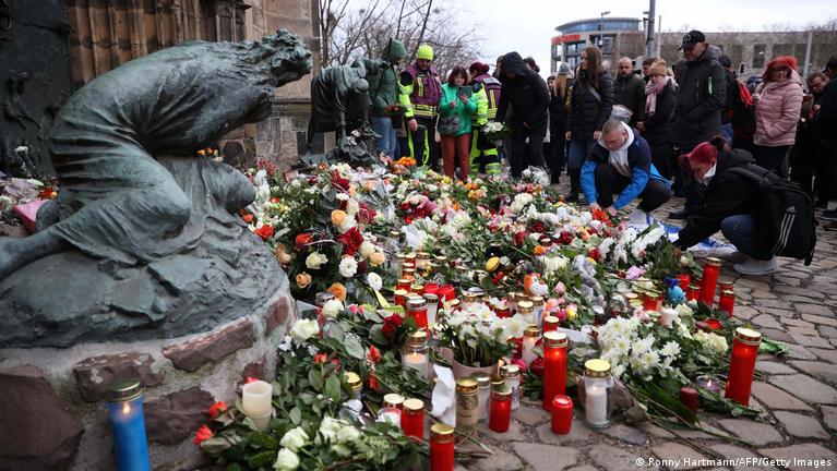 A memorial site with numerous flower bouquets and candles on the ground, surrounded by a crowd of people paying their respects.