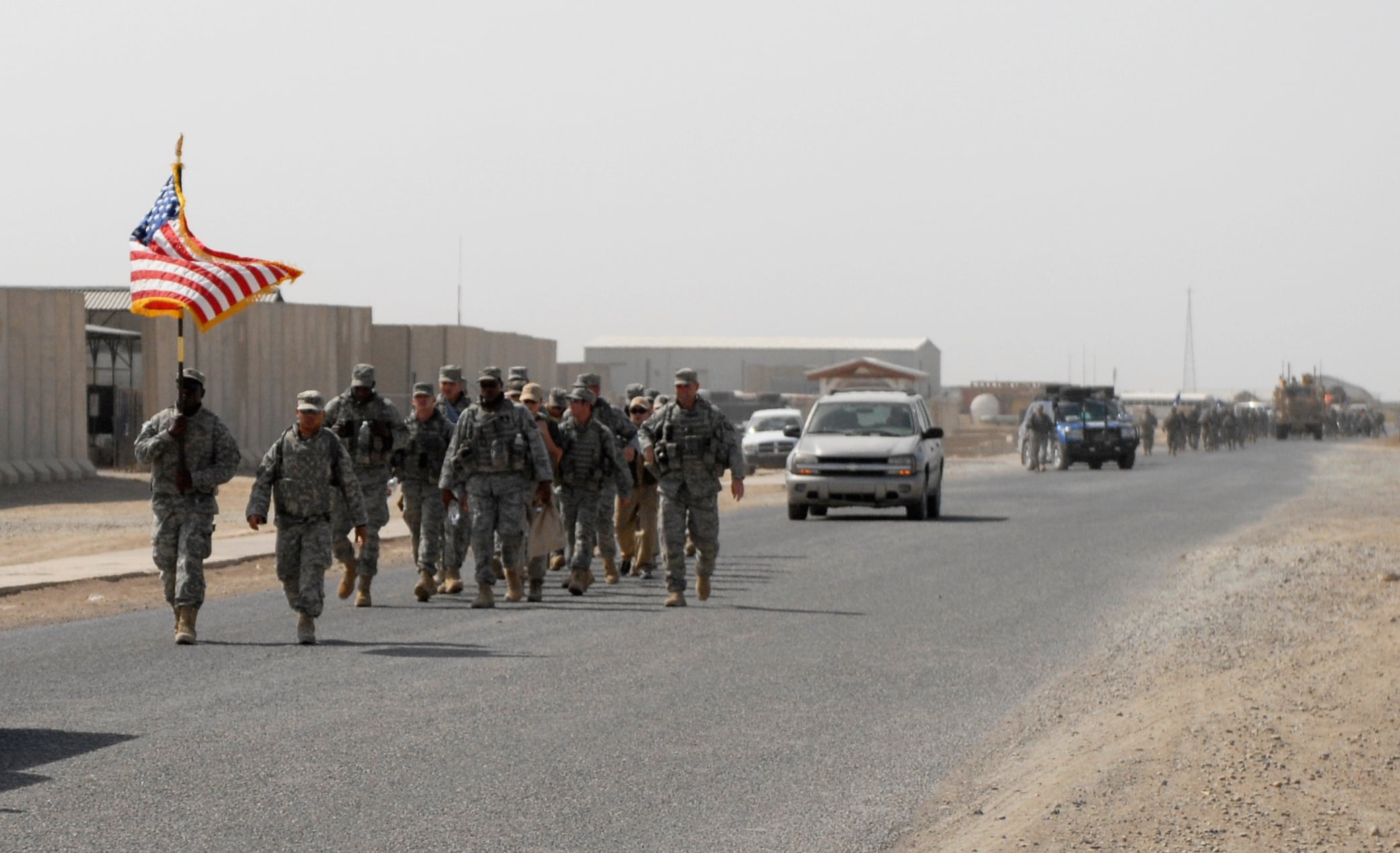 Soldiers in camouflage march on a road with an American flag, vehicles follow, and desert buildings are visible in the background.