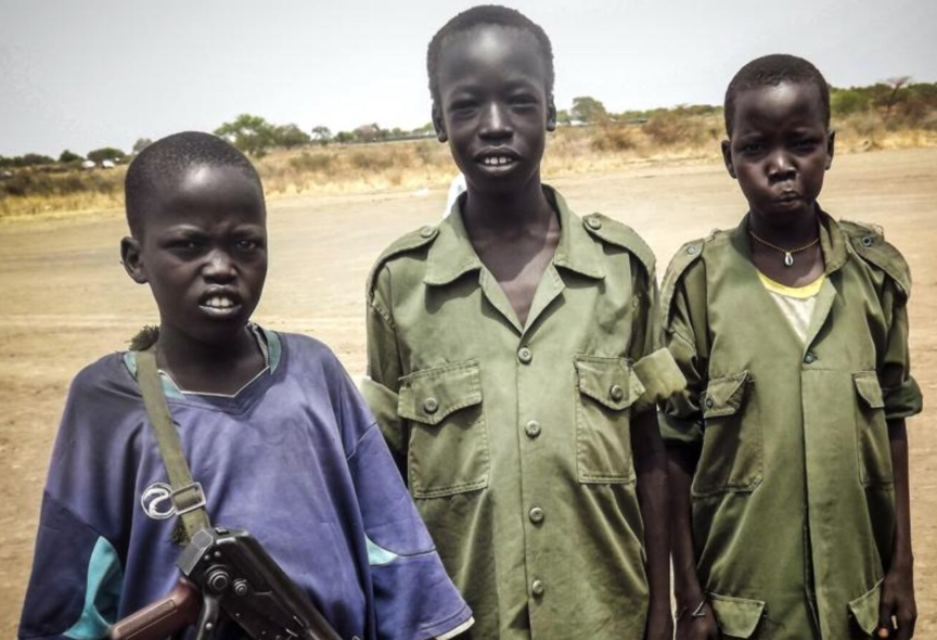 Three young boys in military-style attire stand outdoors on a dirt field. One holds a rifle, and dry grassland is visible in the background.
