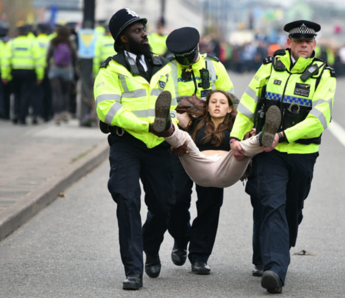 Police officers carry a woman away from a protest scene, with other officers and protesters visible in the background.