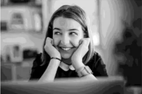Woman smiling with hands on her cheeks, seated indoors. She wears a dark top and a watch, with a blurred background including shelves and books.