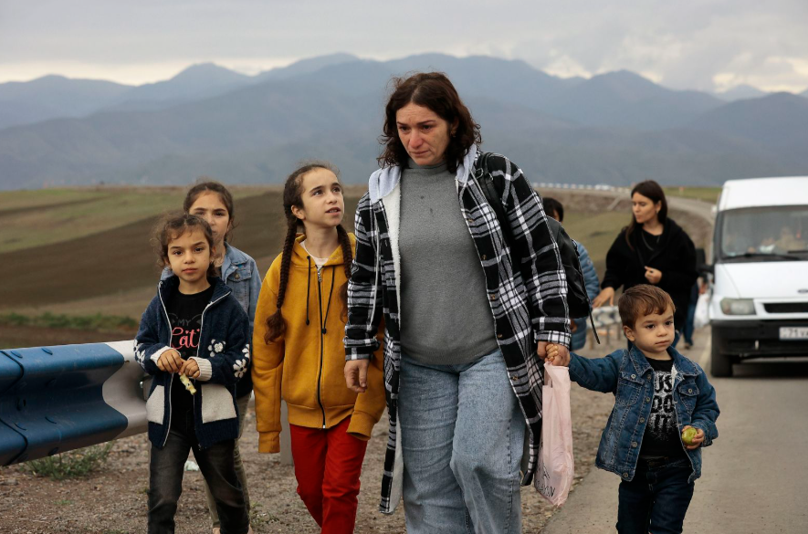 A woman walks with four children in casual clothing on a rural road, with mountains in the background and a white van parked nearby.