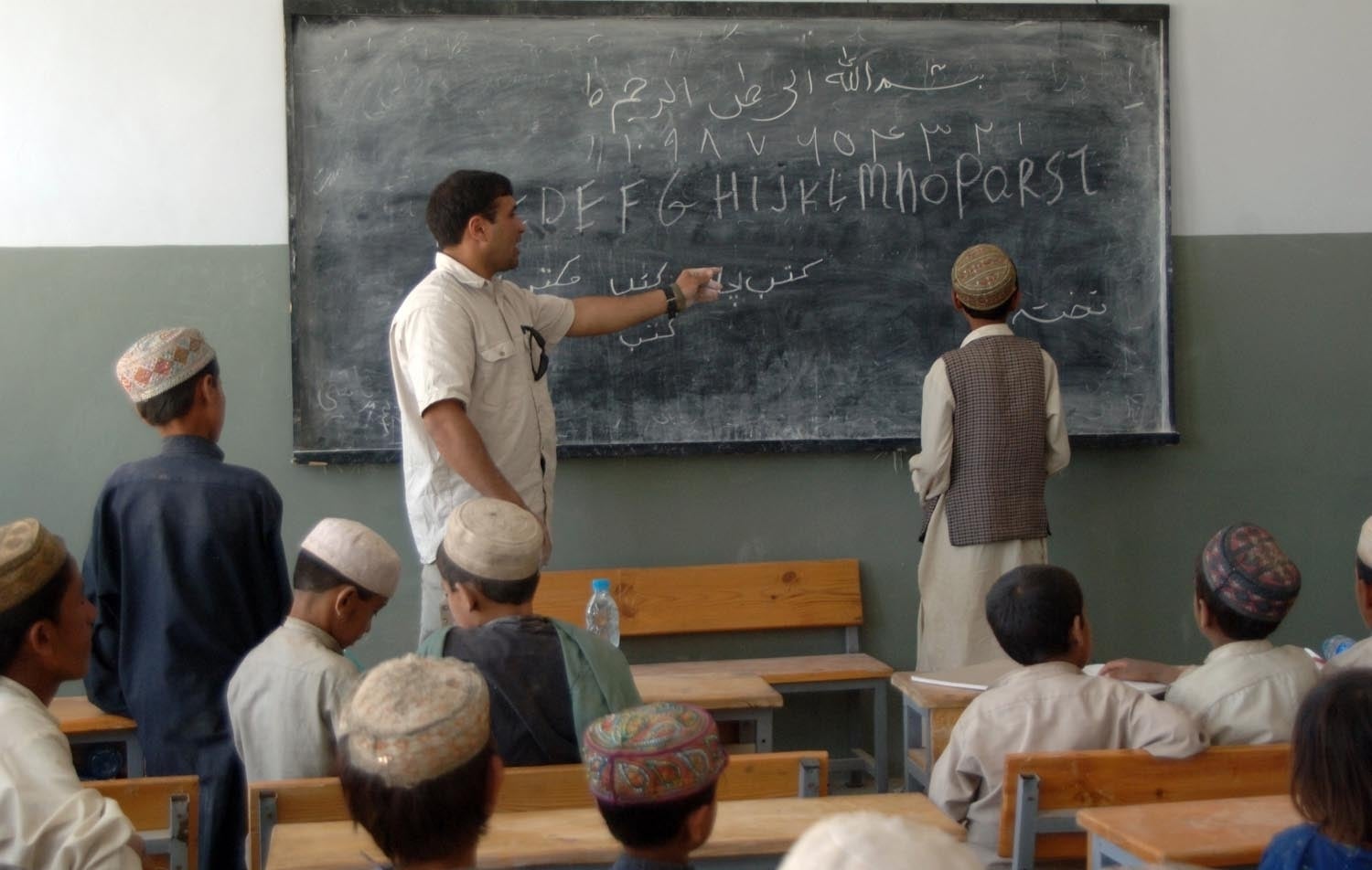 A teacher points to a blackboard with Arabic and English writing, teaching boys in traditional clothing seated at wooden desks.