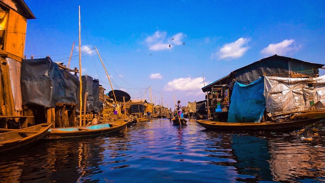 A man paddles a canoe through a narrow waterway lined with wooden shacks covered in tarps and metal under a bright blue sky with scattered clouds.
