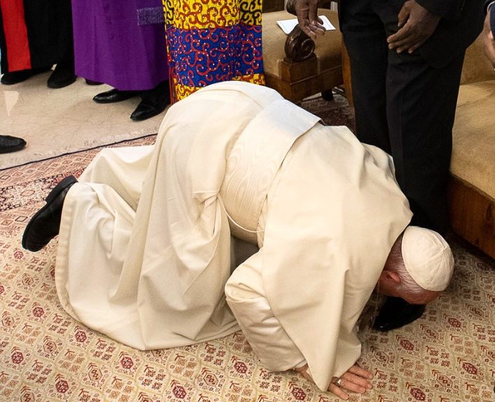 A man in a white robe kneels, kissing the feet of another standing individual, with people in colourful robes in the background.