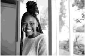 Smiling person with braided hair stands near a window, wearing a light jumper. Sunlight illuminates the room, and trees are visible outside.