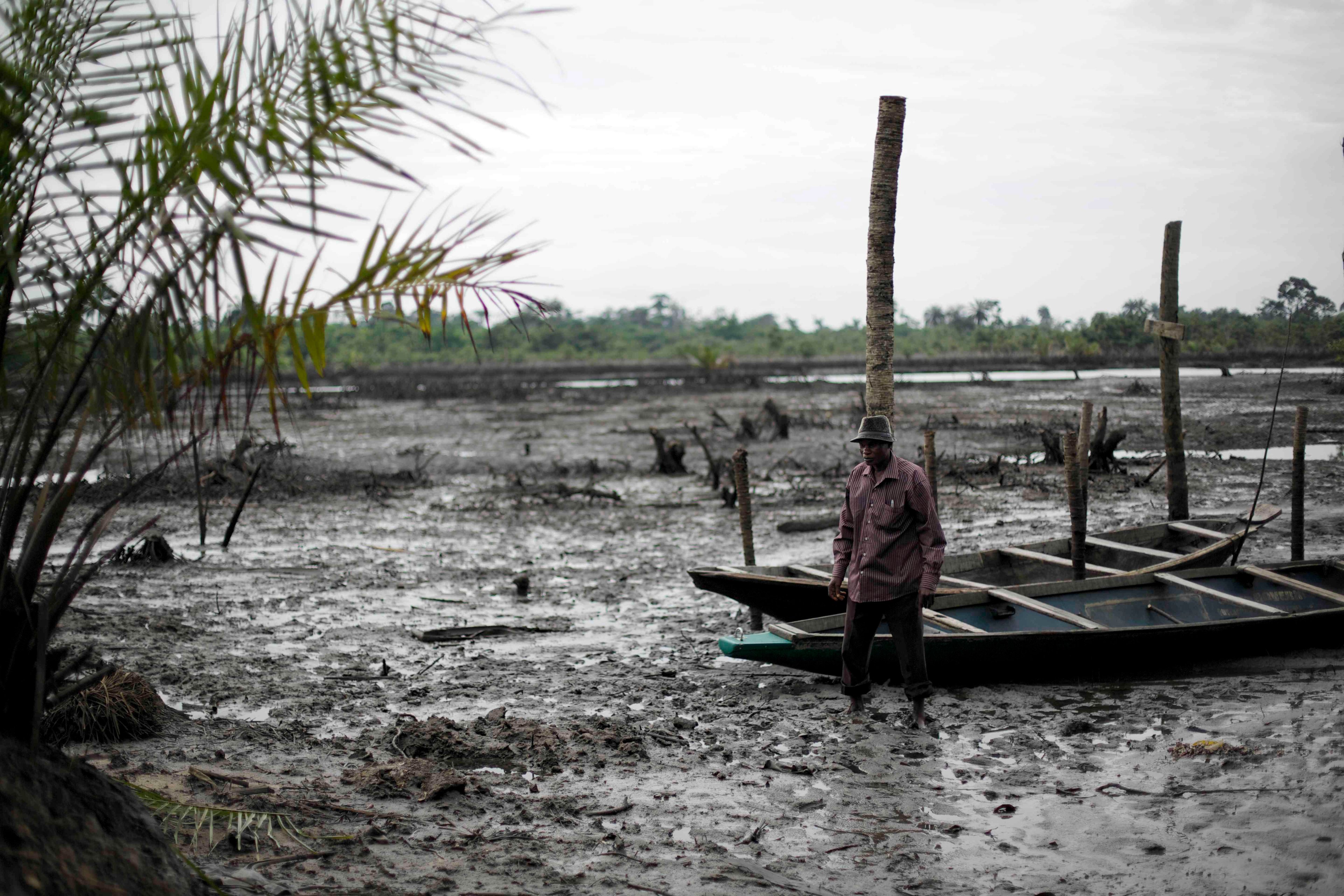 Man in muddy landscape standing by a wooden boat, with sparse vegetation and palm leaves in the foreground under an overcast sky.