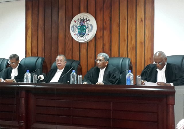 Four judges in black robes sit at a large wooden bench in a courtroom. A coat of arms decorates the wooden panel behind them.