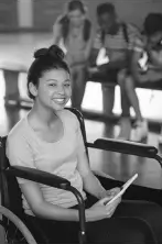 Young woman in a wheelchair smiling, holding a tablet. In the background, three students sit on a bench, engaged with electronic devices.