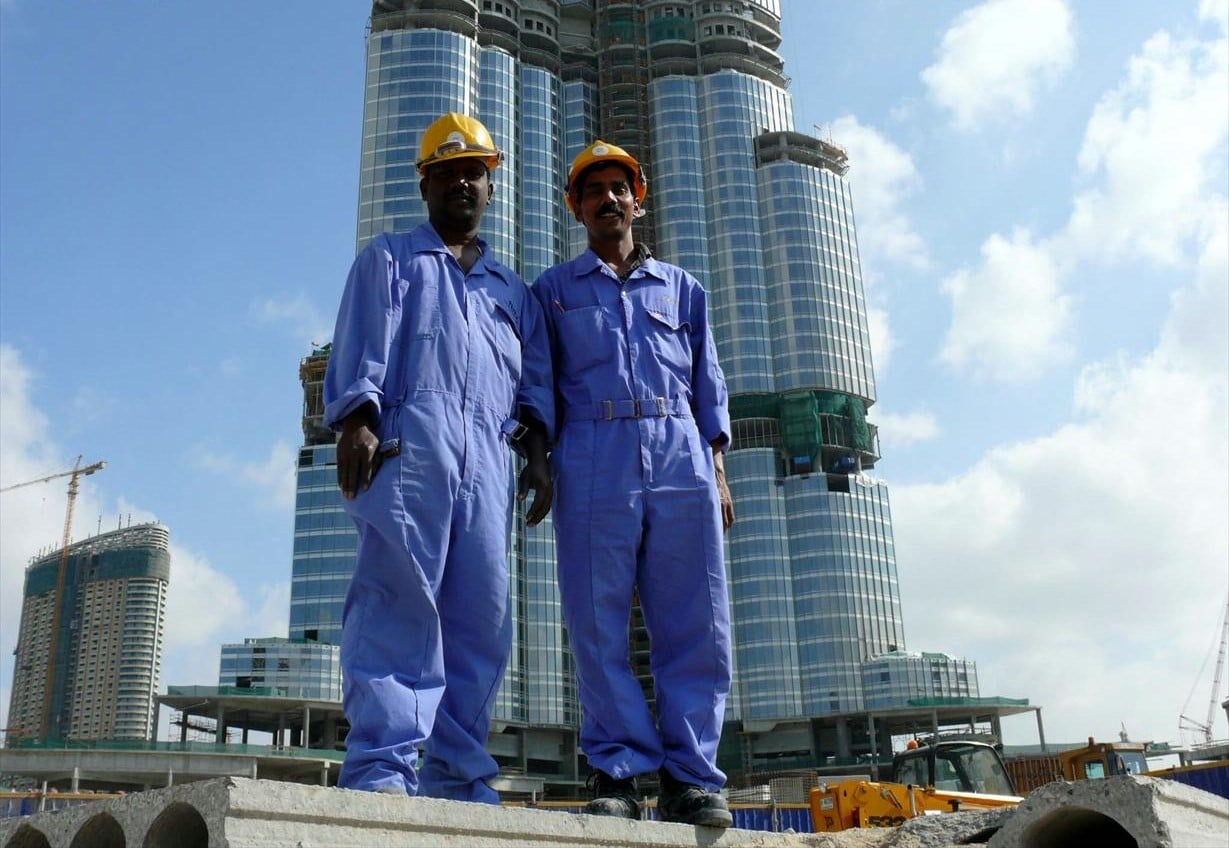 Two construction workers in blue overalls and yellow helmets stand in front of an unfinished skyscraper under a blue sky.