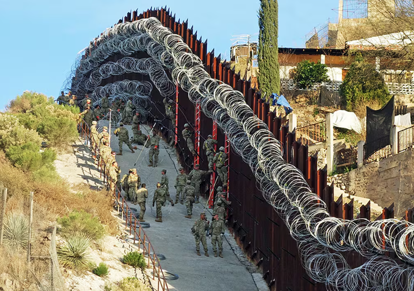 Soldiers in camouflage uniforms reinforce a steep border fence lined with coils of razor wire beside a hillside with sparse vegetation and nearby houses