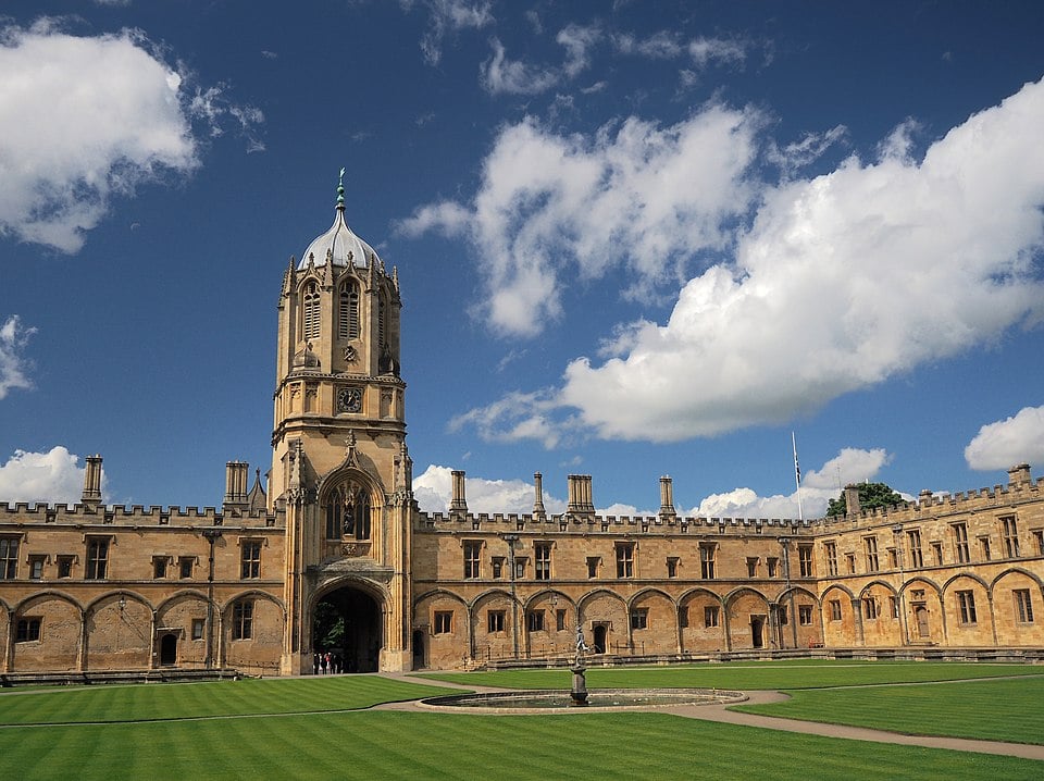 Historic stone building with a clock tower and ornate windows, surrounding a circular lawn with a fountain in the centre, under a cloudy sky.