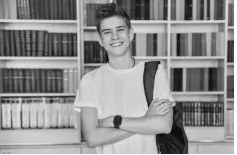 Teenager in a white T-shirt, smiling with crossed arms, stands in front of a bookshelf filled with neatly arranged books, carrying a backpack.