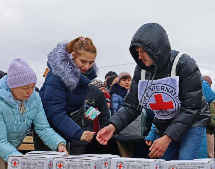 A humanitarian worker in a coat distributes Red Cross aid boxes to a group of people in winter clothing, outside on a cloudy day.