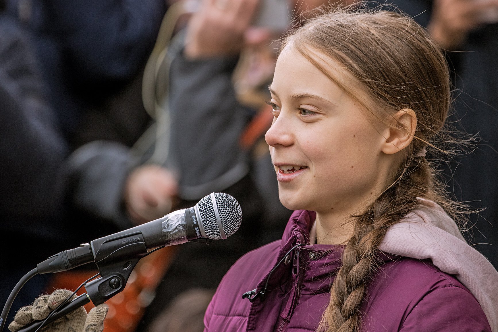 Young girl with braided hair speaking into a microphone outdoors, wearing a purple coat, with a blurred crowd in the background, smiling slightly.