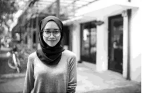 Young woman in a headscarf and glasses smiles while standing on a street. There are buildings and a bicycle in the blurred background. Black and white image.