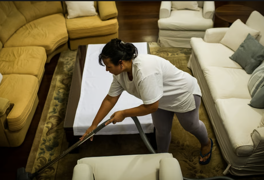 A woman in casual clothing is vacuuming a rug in a living room with beige and white sofas, a wooden coffee table, and a patterned carpet.