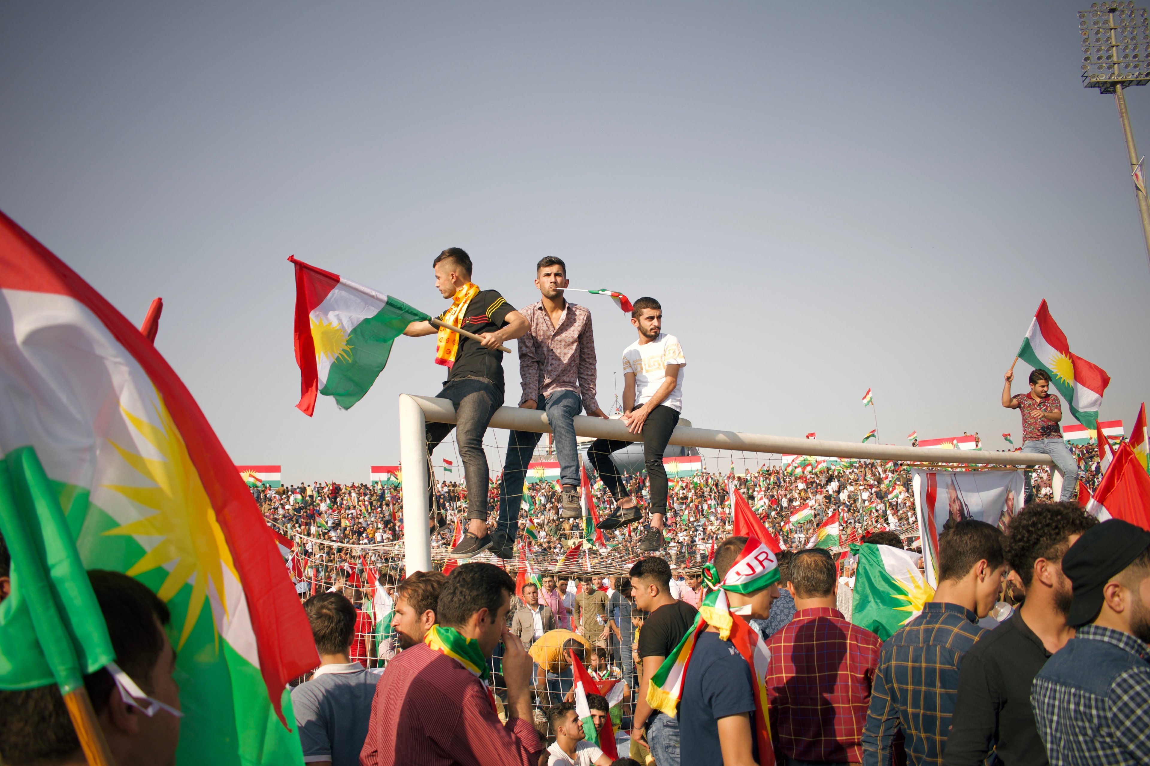 A crowded pro-Kurdistan independence rally at Franso Hariri Stadium, Kurdistan, Iraq, with protestors sitting atop a goalpost and Kurdish flags being waved