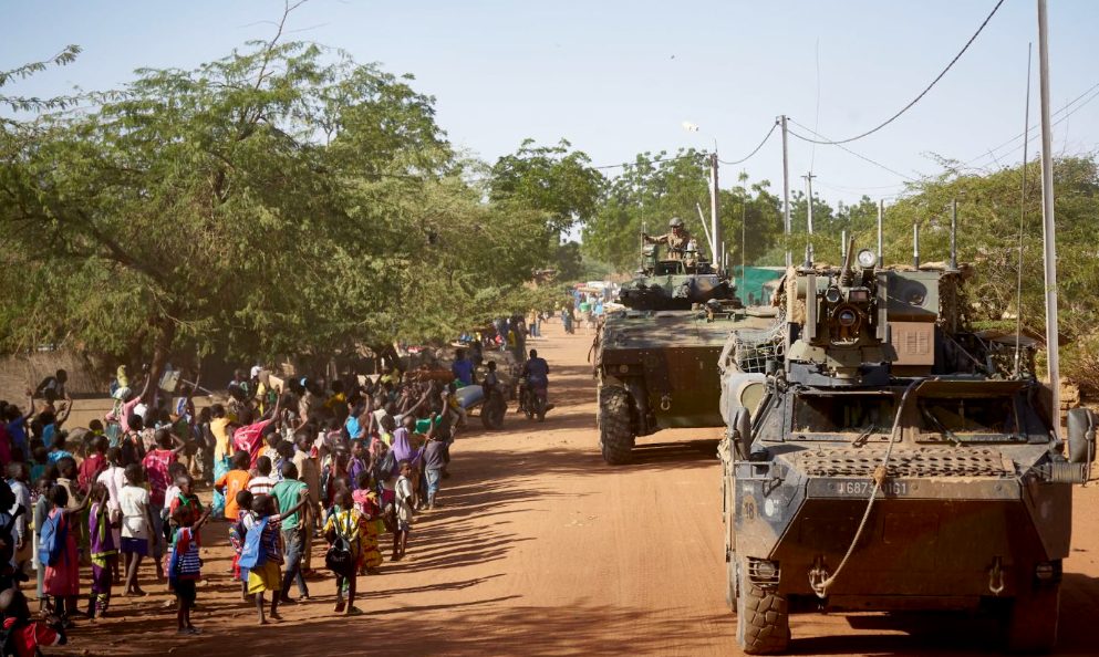 Military vehicles move through a dusty street lined with trees, accompanied by a large crowd of people, including children in colourful clothing.