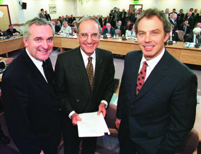 Three men in suits smiling at a conference room. The central man holds documents. Attendees and media equipment are visible in the background.
