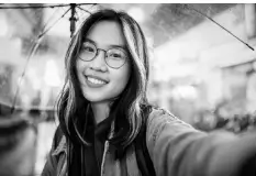 Young woman smiling under a transparent umbrella in a street setting, wearing glasses and a casual jacket, in a black and white photograph.