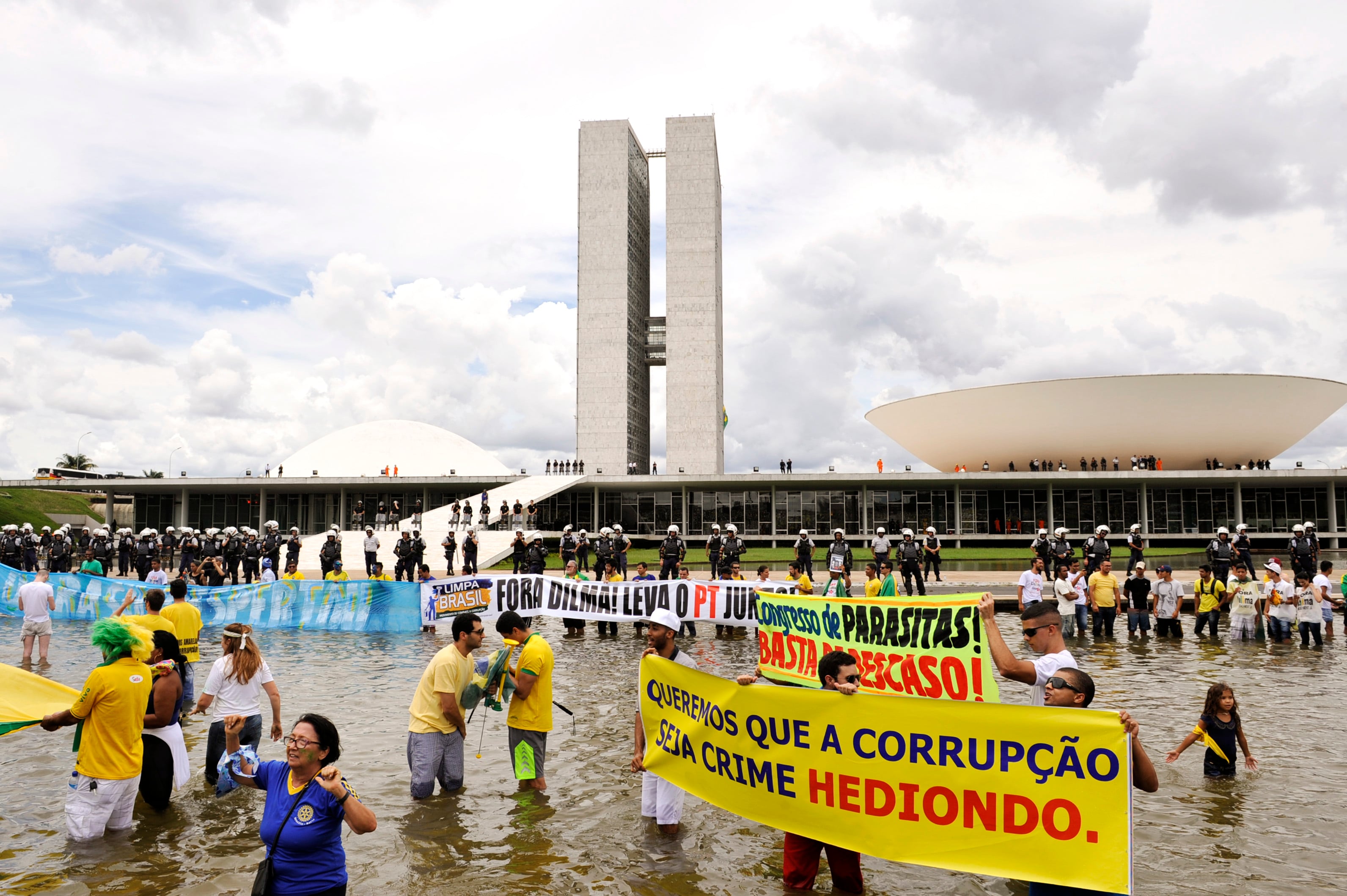 Protesters in water hold banners against corruption in front of a government building, guarded by police, with a cloudy sky overhead.