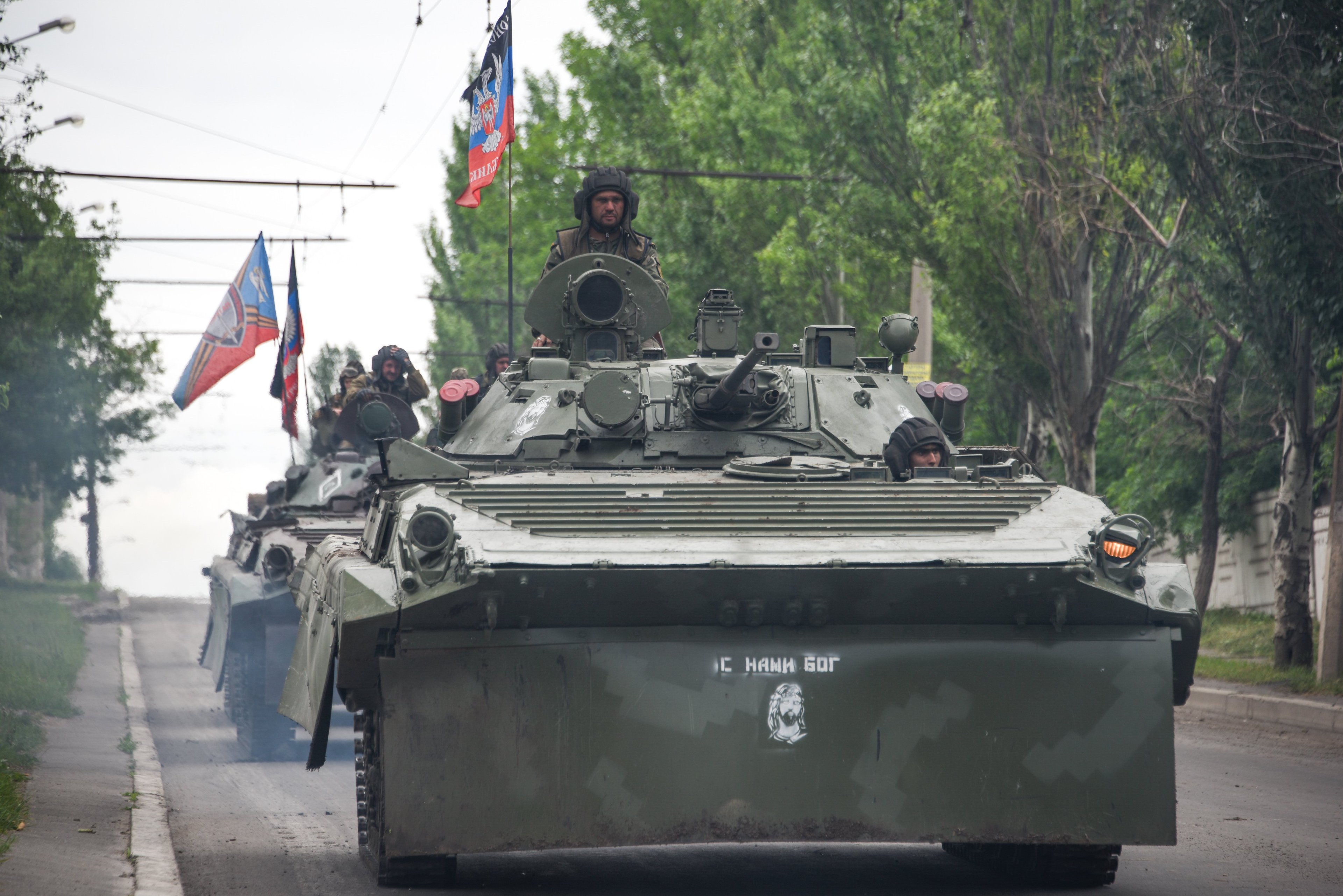A series of tanks with flags drive along a tree-lined street, with helmeted soldiers visible in tank turrets