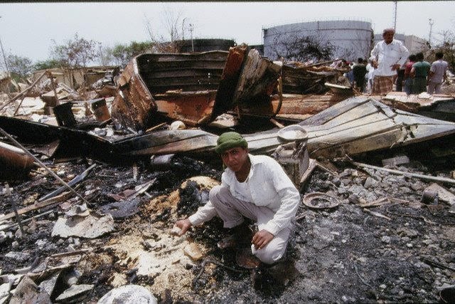Man crouching amid charred debris and wreckage in an outdoor setting, with scattered remains and large tanks in the background.