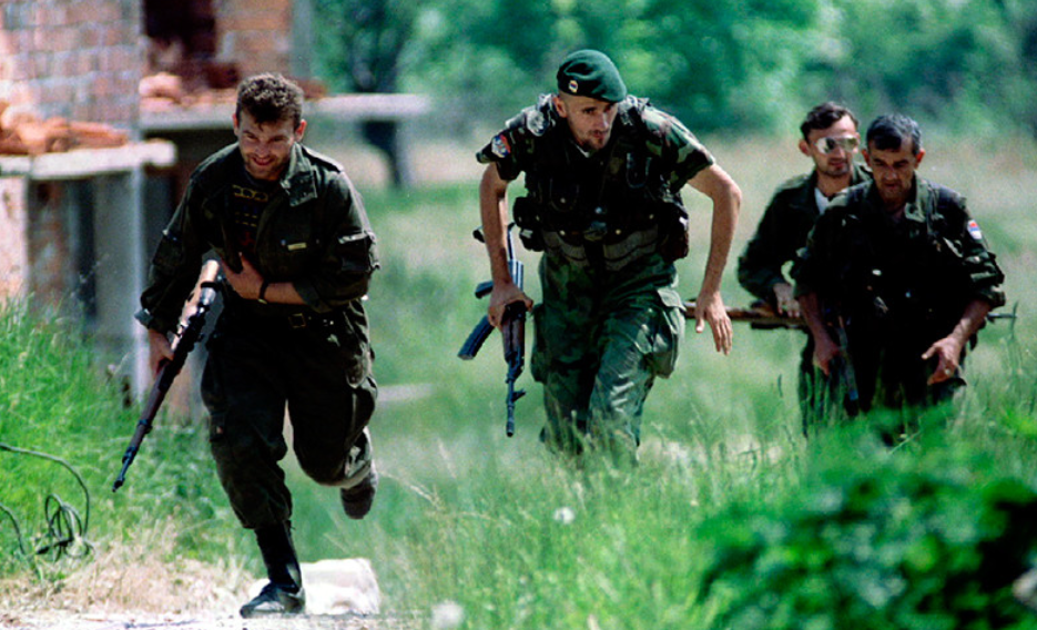Four soldiers in camouflage uniforms run through grassy terrain next to a brick building, carrying rifles, with trees in the background.