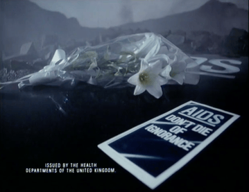 White lilies wrapped in plastic lie on a dark surface beside a leaflet reading “AIDS: Don’t Die of Ignorance”, with UK health department text below.