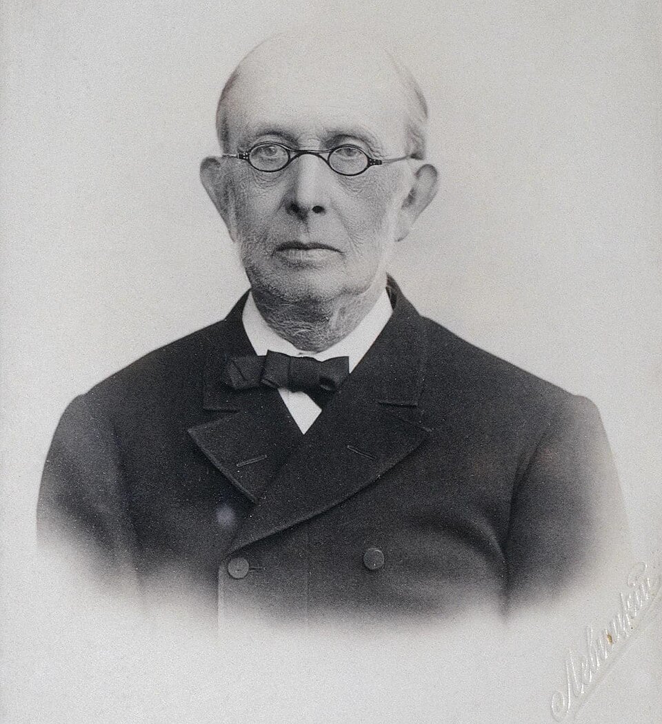 Old black-and-white studio portrait of a serious elderly man in round spectacles and dark suit with bow tie, facing the camera against a plain background